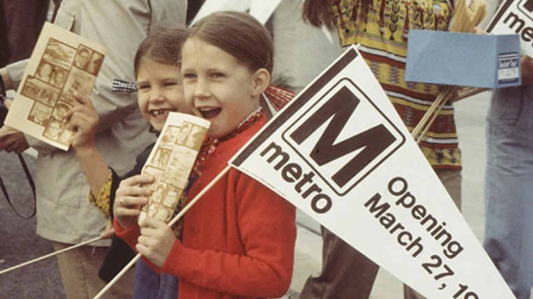 young girl in red sweather holding a banner that says 'Metro opening March 27th'