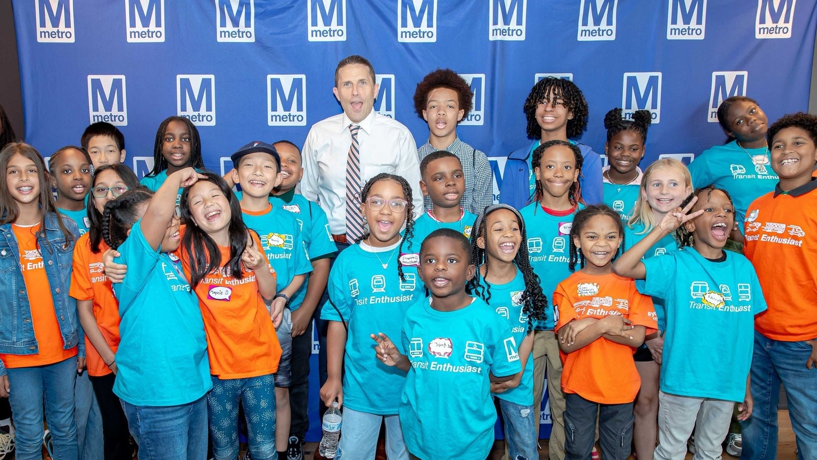 Metro General Manager Randy Clarke poses with children wearing Transit Enthusiast shirts in front of a Metro backdrop