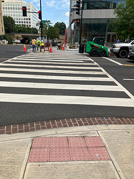A picture of the crosswalk across D St at its intersection with 7th St. This crosswalk leads to the WMATA Headquarters. There are construction workers in the picture that were at the intersection that day.