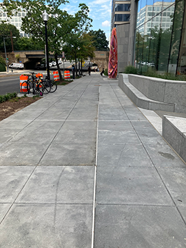 A picture of a sidewalk down 7th Street. The WMATA Headquarters building is on the left, and the street is on the right. There is landscaping and some construction cones to the right before the street. A picture of a sidewalk down 7th Street. The WMATA Headquarters building is on the left, and the street is on the right. There is landscaping and some construction cones to the right before the street.