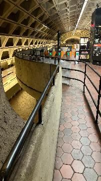 A picture of the guide rails to the faregates. The faregates are in the background. To the right is the railing and to the left is a half wall with railing on top of it.
