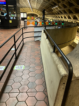 A picture of the guide rail pathing towards the faregates on the mezzanine level. The railing is on the left and there is a concrete wall on the right.