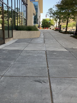 A picture of the sidewalk along 7th Street. The WMATA HQ building is on the left and 7th Street is on the right. There is landscaping in the right of the picture.