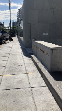 A picture of the barriers directly in front of the Transit Accessibility Center. There is a parapet wall to the right on a raised ledge. Beyond it is a bollard with space on either side between it, the parapet wall, and the building to pass through.