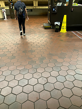 A picture of the walkway past the station manager's kiosk to the ADA faregate. In the background and to the right the station manager's kiosk has a yellow wet floor cone in front of it and to the left there is a man walking away from the camera.